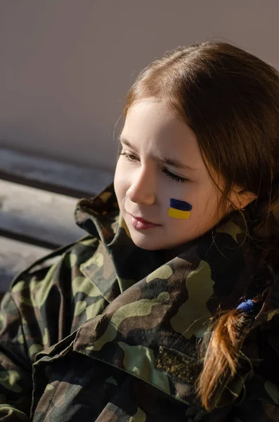 Criança triste com bandeira ucraniana pintada na bochecha e casaco de camuflagem — Fotografia de Stock