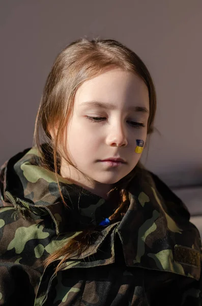 Worried kid with painted ukrainian flag on cheek and camouflage jacket — Stock Photo