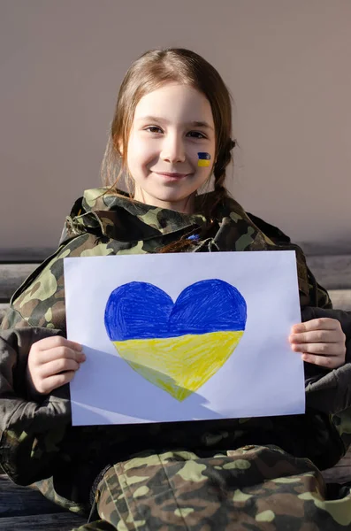 Fröhliches Kind mit bemalter ukrainischer Flagge auf der Wange und Tarnjacke mit Papier mit Herz und Nationalflaggenfarben — Stockfoto