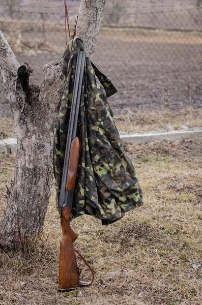 Camouflage jacket hanging on tree near rifle in woods — Stock Photo