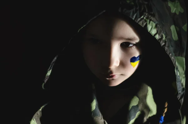 Niño triste con la bandera pintada de Ucrania en la mejilla con camuflaje capucha aislado en negro - foto de stock