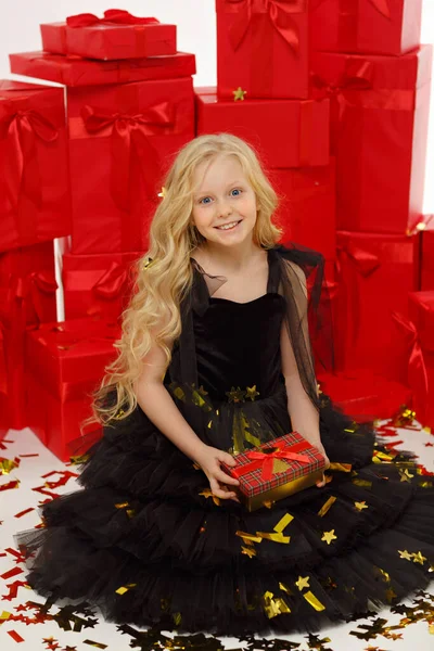 黒のドレスの魅力的なブロンドの女の子は 新年のための贈り物を保持し 赤の贈り物とコンフェッティの背景に座っています ストック画像