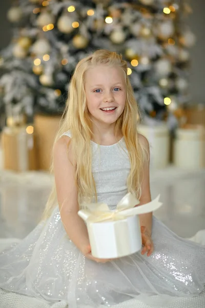 クリスマスツリーの近くの銀のドレスの魅力的な小さなブロンドの女の子は 感情的に贈り物に驚いています ロイヤリティフリーのストック画像