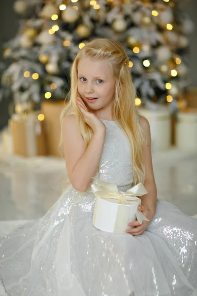 クリスマスツリーの近くの銀のドレスの魅力的な小さなブロンドの女の子は 感情的に贈り物に驚いています ストック画像