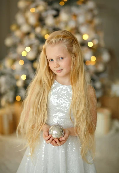 クリスマスツリーの近くの銀のドレスの魅力的な小さなブロンドの女の子は彼女の手の中におもちゃを持っています ロイヤリティフリーのストック画像
