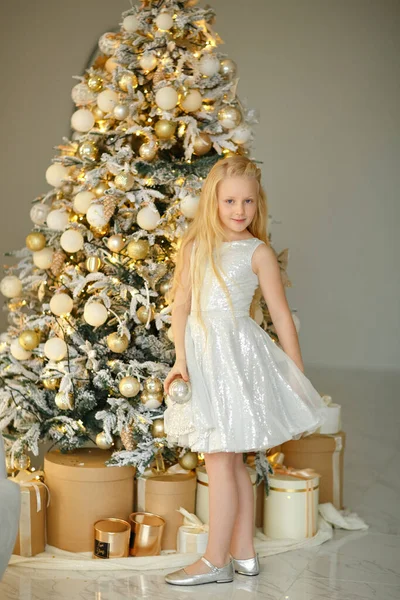 銀のドレスの魅力的な小さなブロンドの女の子は 内部のクリスマスツリーの近くに立っています ロイヤリティフリーのストック写真