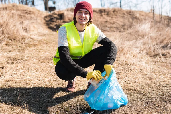 Man nature activist in yellow vest smiles and crouches down to collect some trash and put it into blue trash bag