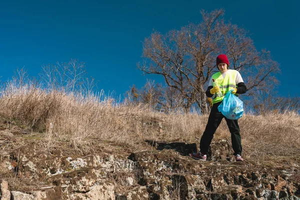 Man nature activist in yellow vest puts glass bottle in the blue trash bag outdoors