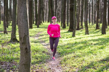 Gül rengi elbiseler giyen genç bir kadın patika boyunca güneşli bir bahar ormanında koşuyor.