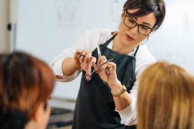 Kuaförün elleri kuaför makası tutuyor, öğrencilere saç kesme tekniklerini anlatıyor.