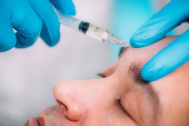 Erkekler için yaşlanma karşıtı tedaviler. Ellerinde cerrahi eldiven var. Deri doldurucu enjekte ediyorlar. Orta yaşlı erkeklerin yüzü...   