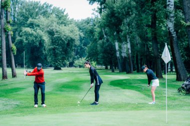 Golf sahasında golf hocasıyla atış talimi yapan iki genç golfçü.