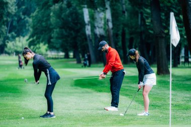 Golf sahasında golf hocasıyla atış talimi yapan iki genç golfçü.