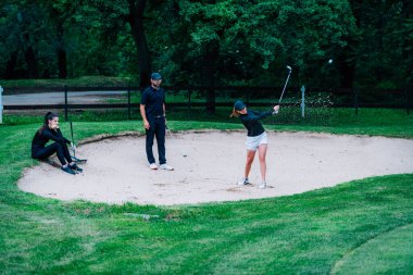 Golf dersleri alan genç bir kadın golf dersi alıyor, kum deposundan oynuyor. 