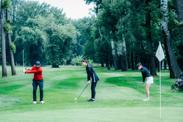 Golf sahasında golf hocasıyla atış talimi yapan iki genç golfçü.