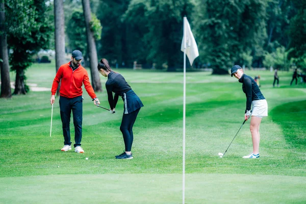 Golf sahasında golf hocasıyla atış talimi yapan iki genç golfçü.