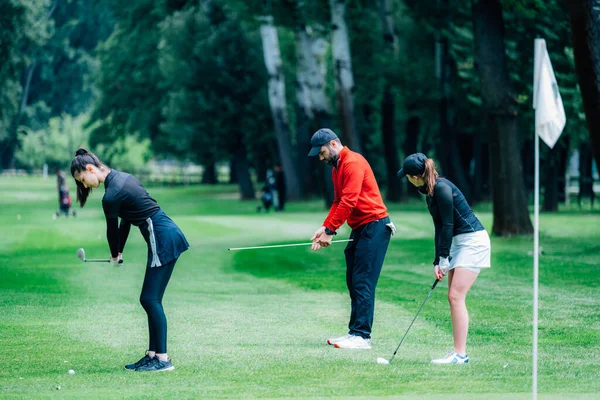 Golf sahasında golf hocasıyla atış talimi yapan iki genç golfçü.