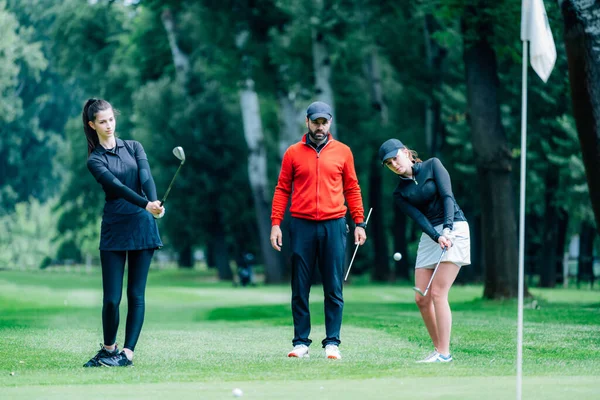 Golf sahasında golf hocasıyla atış talimi yapan iki genç golfçü.