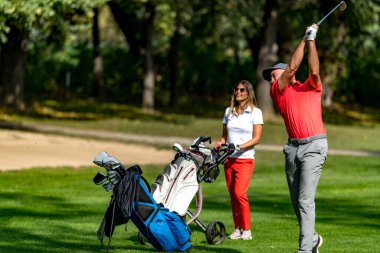 Golf sahasında golf oynayan bir çift, güneşli bir günde golf oynuyorlar, adam topa vuruyor.