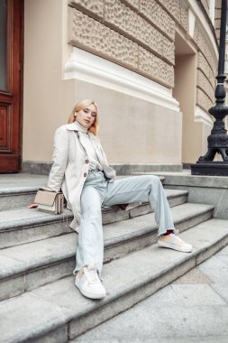 Trençkotlu genç moda sarışın kadın şehrin merdivenlerinde oturuyor.
