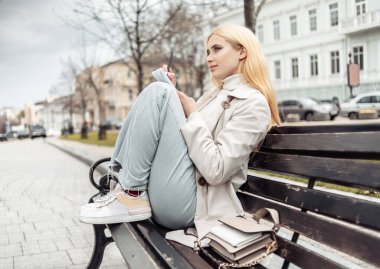 Tarz sahibi genç bir kadın bir bankta oturur ve defterine yazar.