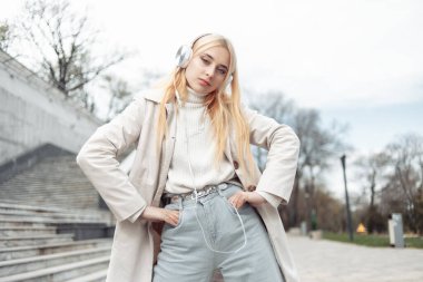 Trençkotlu genç, şık sarışın kadın dışarıda kulaklıkla müzik dinliyor.