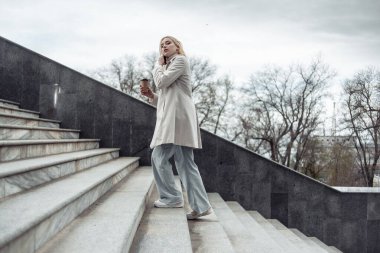 Elinde kahve fincanı olan genç sarışın kız merdivenlerden yukarı çıkarken telefonla konuşuyor.