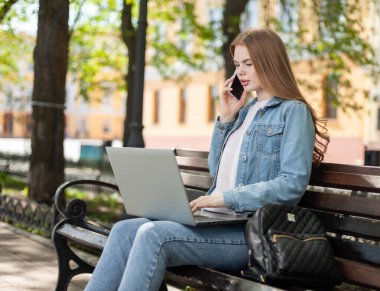 Genç, güzel bir kız öğrenci ya da serbest çalışan, dizüstü bilgisayar kullanıyor ve şehirde bir bankta otururken telefonla konuşuyor.