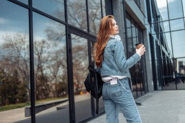 Kot ceketli ve iş merkezinin penceresinin yanında kahve fincanı tutan güzel bir kadın.