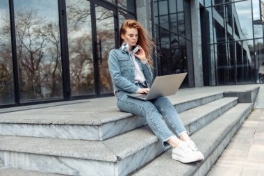 Genç bir kız öğrenci ya da serbest çalışan elinde dizüstü bilgisayarla merdivenlerde oturuyor.