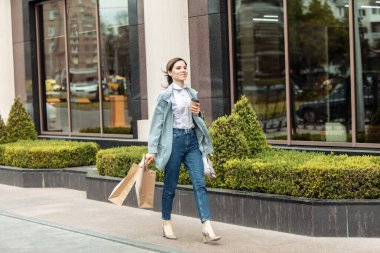 Güzel, neşeli bayan alışveriş torbalarıyla şehir caddesinde yürüyor ve yolda kahve içiyor. Yaşam tarzı alışveriş konsepti