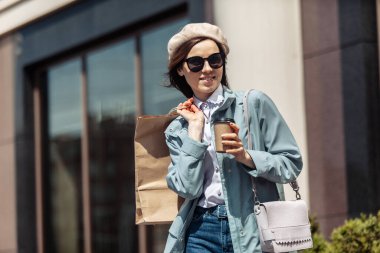 Moda trençkotlu, bereli şık alışveriş meraklısı bir kadın, alışveriş torbaları ve bir fincan kahveyle şehirde güneş gözlüğü. Alışveriş konsepti. Yaşam biçimi