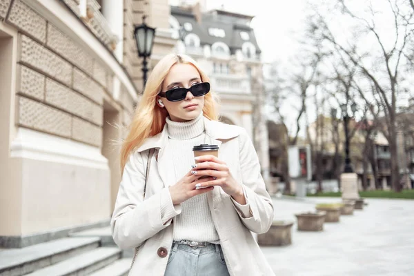 年轻的金发女郎 戴着太阳镜 穿着风衣 一边走一边拿着一杯咖啡 — 图库照片