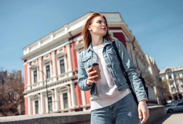 Beautiful woman in a denim jacket drinks coffee on the go in city