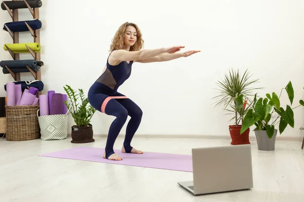 Young attractive caucasian woman exercising with fitness rubber bands watching online workout on laptop in yoga studio