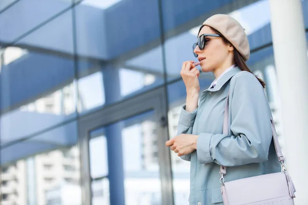 サングラスとベレー帽の若いスタイリッシュな女性は ビジネスセンターの窓の背景に彼女の唇を描きます ファッション 美しさ ライフスタイル — ストック写真
