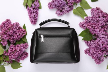 Beyaz arka planda çiçek açan leylak dalları olan siyah deri bir çanta. Bahar, moda konsepti. Üst Manzara. Düz yatıyordu