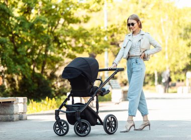Güneş gözlüklü şık bir anne parkta bebek arabasıyla yürüyor.