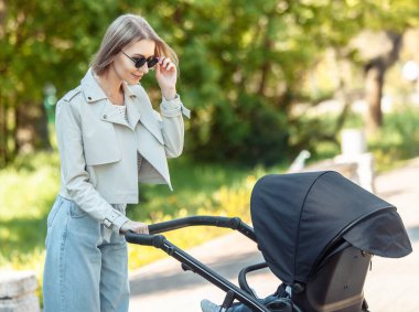 Güneş gözlüklü şık bir anne parkta bebek arabasıyla yürüyor.