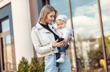 İş dünyasından modern bir anne kızını kollarında tutuyor ve şehir merkezindeki bir akıllı telefona bakıyor. Yaşam biçimi