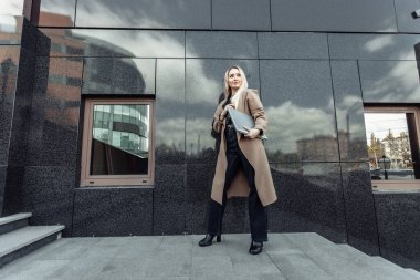 Şehir merkezinde trençkotlu dizüstü bilgisayarı olan şık sarışın iş kadını.