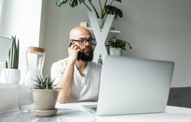 Laptop kullanan sakallı adam, klavyede yazan, e-posta ya da mesaj yazan, telefonla konuşan, bilgisayar üzerinde çalışan, online alışveriş yapan, modern odada oturan başarılı bir serbest yazar.