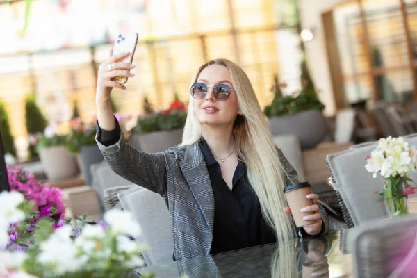 年轻的金发女郎戴着太阳镜 坐在咖啡馆的阳台上 一边喝着咖啡 一边自作主张 — 图库照片