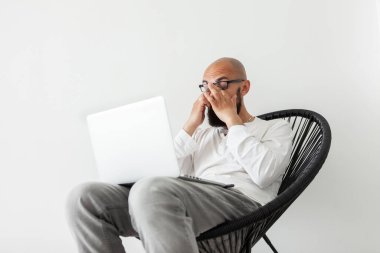 Koltuğunda dizüstü bilgisayarla oturan yorgun sakallı serbest çalışan erkek elleriyle gözlerini ovuyor..