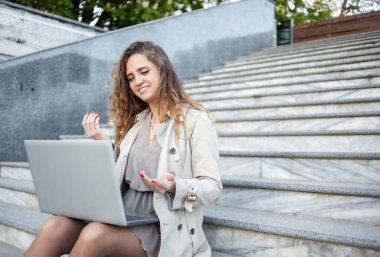 Genç kız öğrencinin dışarıda merdivenlerde otururken bilgisayarında görüntülü arama var.