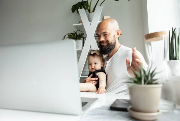 Working father waves to the laptop camera with his little daughter in his arms in a modern room.