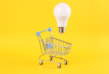 Sarı parlak arka planda yüzen ampullü alışveriş arabası. Asgari alışveriş kavramı