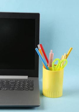 Laptop ve renkli kalemler, okul için çeşitli renkli kağıtlar, mavi arka planda tutucu veya bardak. Uzaklık öğrenme kavramı