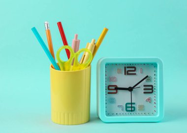 Okul kırtasiyeli plastik bardak, çizim araçları ve mavi arkaplanda alarm saati.