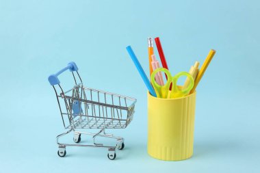 Okul kırtasiyeli plastik bardak, mavi arka planda çizim aletleri ve alışveriş arabası. Okula geri dön.